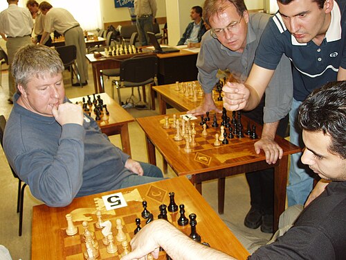 A picture of four men looking over a chessboard and gesticulating. In the background are other chess boards on tables, set up differently.